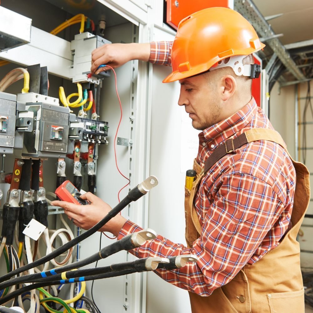 Electrician in orange hard hat and plaid shirt using a multimeter to test electrical panel wires in a commercial facility.